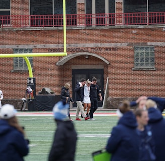 A group of people on a sports field, with some dressed in athletic gear, and others in casual clothing. A brick building labeled 'Gosset Football Team House' is in the background. One of the athletes appears to be aided by others, suggesting a possible injury.