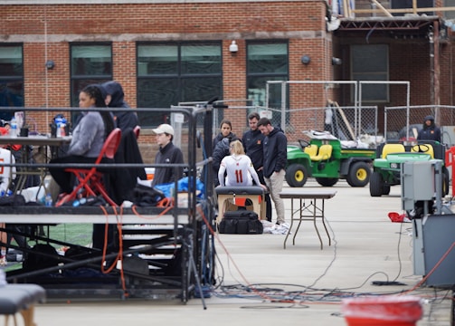 A group of people are gathered around an individual wearing a sports jersey with the number 11. They appear to be in a sports facility area, possibly a training or medical setup. There are tables, chairs, and equipment scattered around, with a brick building in the background. Some individuals are seated on a platform with electronics close by, suggesting a recording or monitoring activity. Utility vehicles and an orange barrier are present in the background.