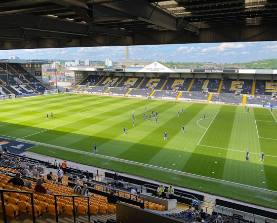 A sports stadium with a well-maintained grass field is depicted. Players are spread across the field, engaging in warm-up activities. The stands are partially filled with spectators, and the stadium is surrounded by urban buildings under a partly cloudy sky.