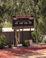 An outdoor scoreboard for San Francisco State is seen among trees and greenery. The board displays sections for shots on goal and corner kicks, with areas for both home and guest teams. It is situated near a running track with a building in the background.